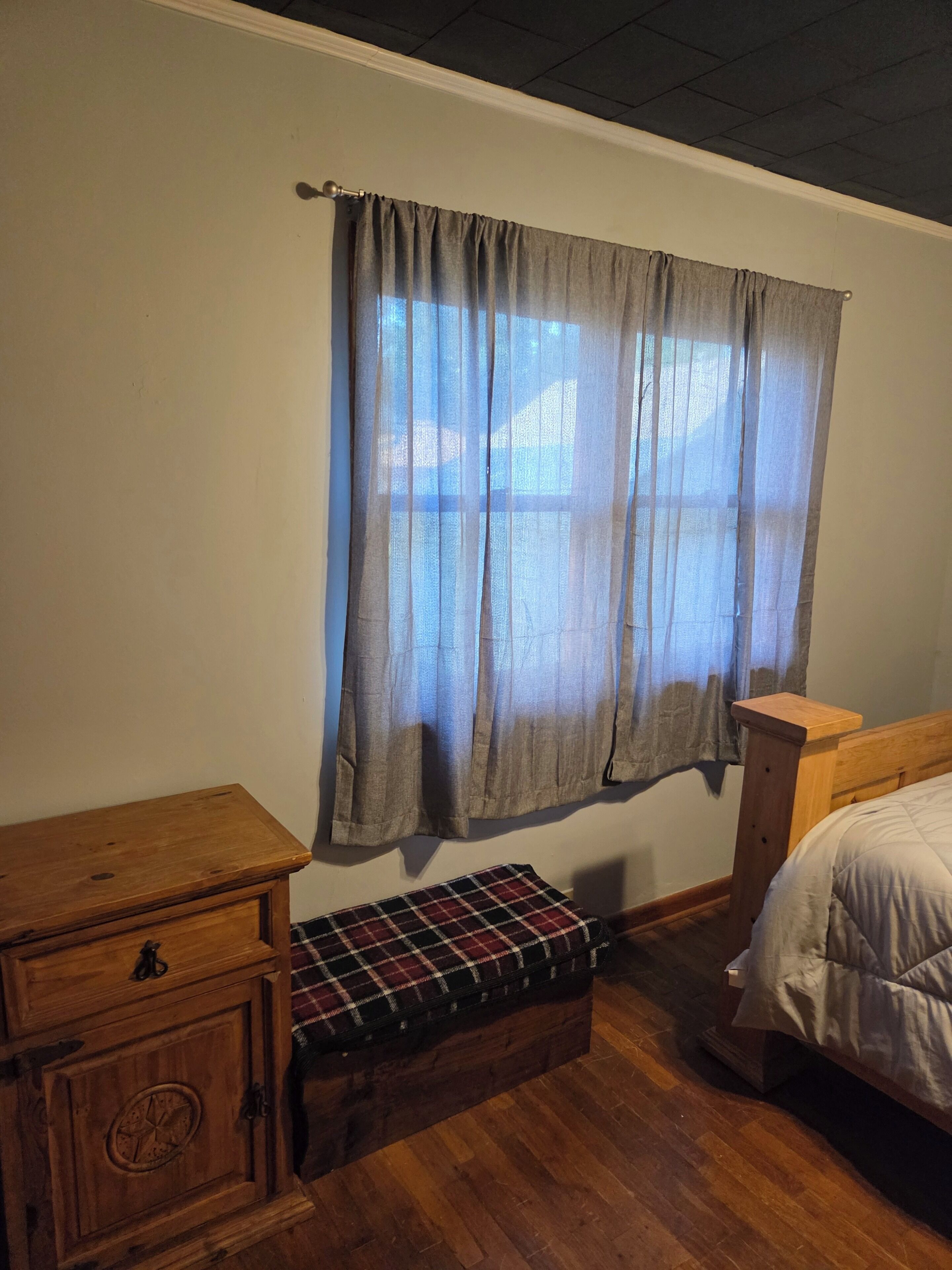 Une chambre à coucher avec une table de chevet en bois, un banc à carreaux, un lit et une fenêtre avec des rideaux.