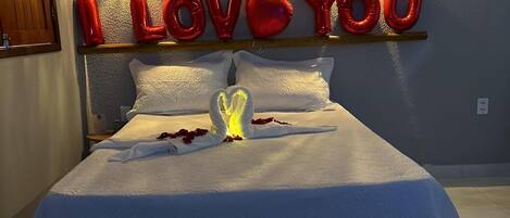 A bed with a white bedspread, a heart-shaped decoration, and red balloons spelling 'I LOVE YOU' on the wall.