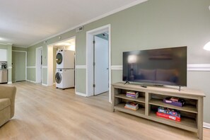 A living room with a flat-screen TV, a sofa, a wooden entertainment center with books, and a laundry area with a washing machine and dryer. - 1 Mi to Beach! HHI Extended Escape w/ Balcony (Hilton Head Island)