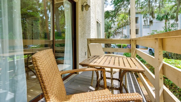 Une terrasse en bois avec des meubles en osier, une porte vitrée et une vue sur la verdure et les bâtiments.