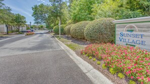 Un panneau de signalisation indiquant l’entrée de Sunset Village, avec une route bordée d’arbres et de fleurs.