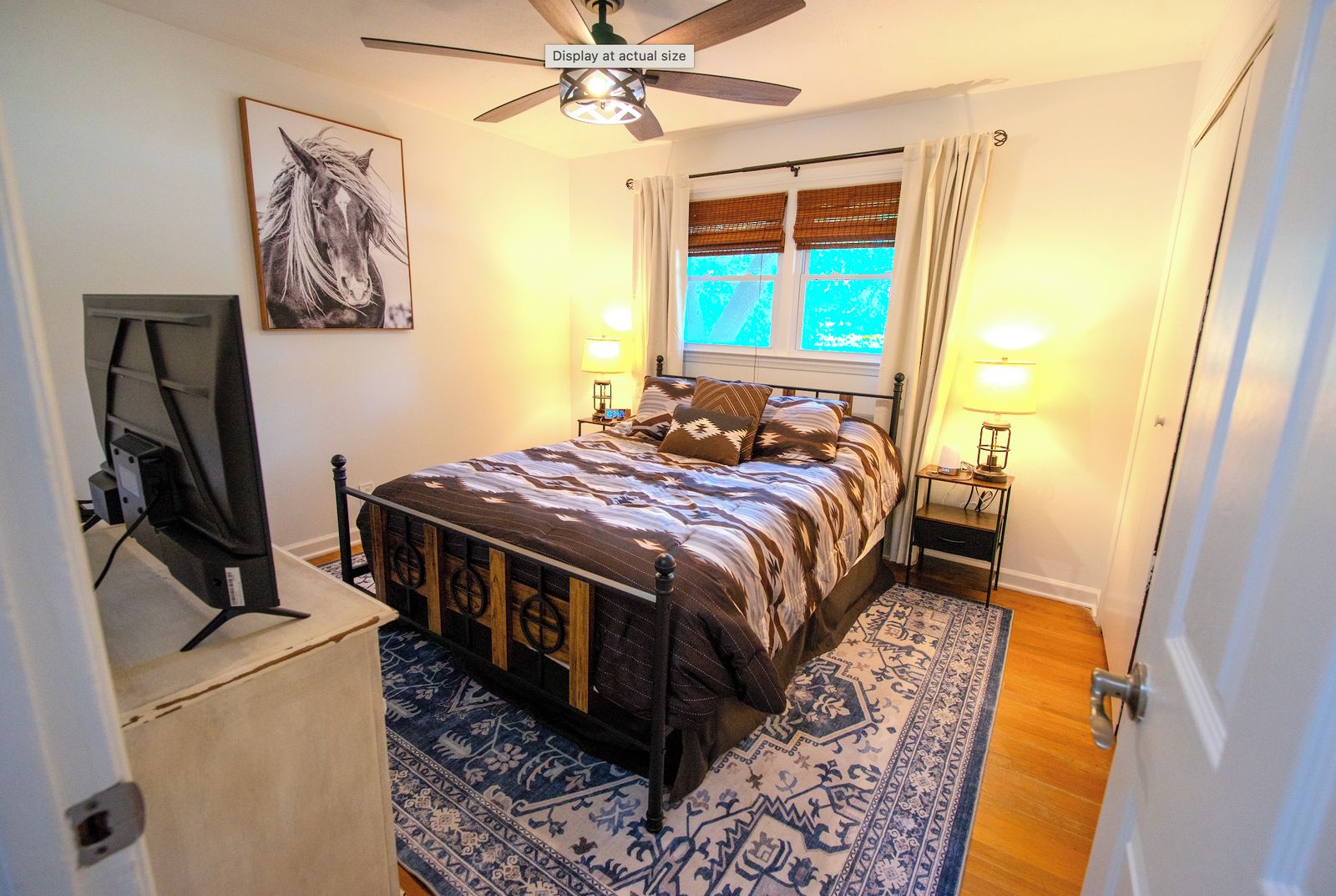 A bedroom with a bed, a ceiling fan, a window with curtains, a nightstand with a lamp, and a wall-mounted TV.