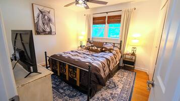 A bedroom with a bed, a ceiling fan, a window with curtains, a nightstand with a lamp, and a wall-mounted TV.