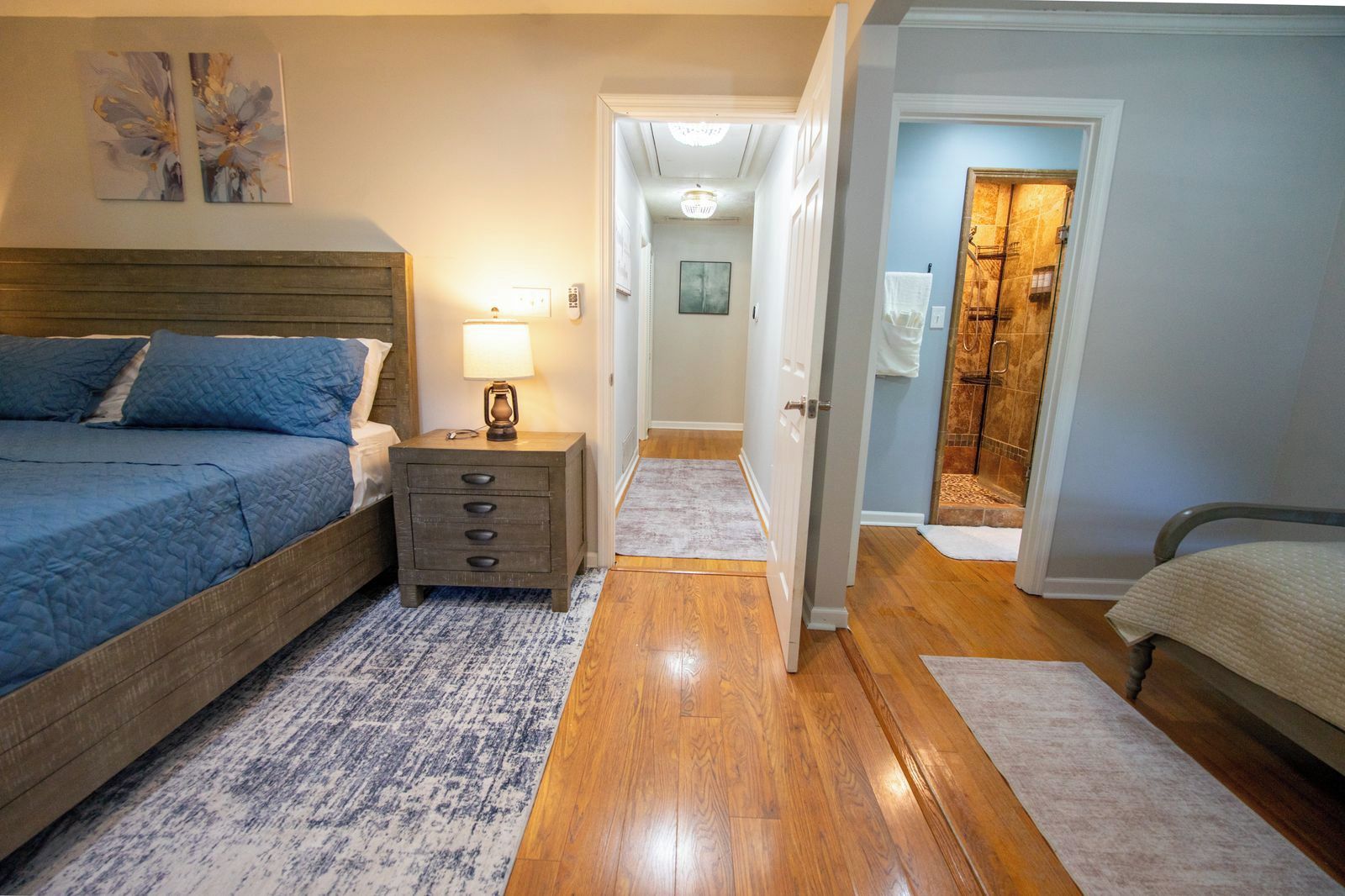 A bedroom with a bed, bedside table, lamp, and a hallway leading to a bathroom.