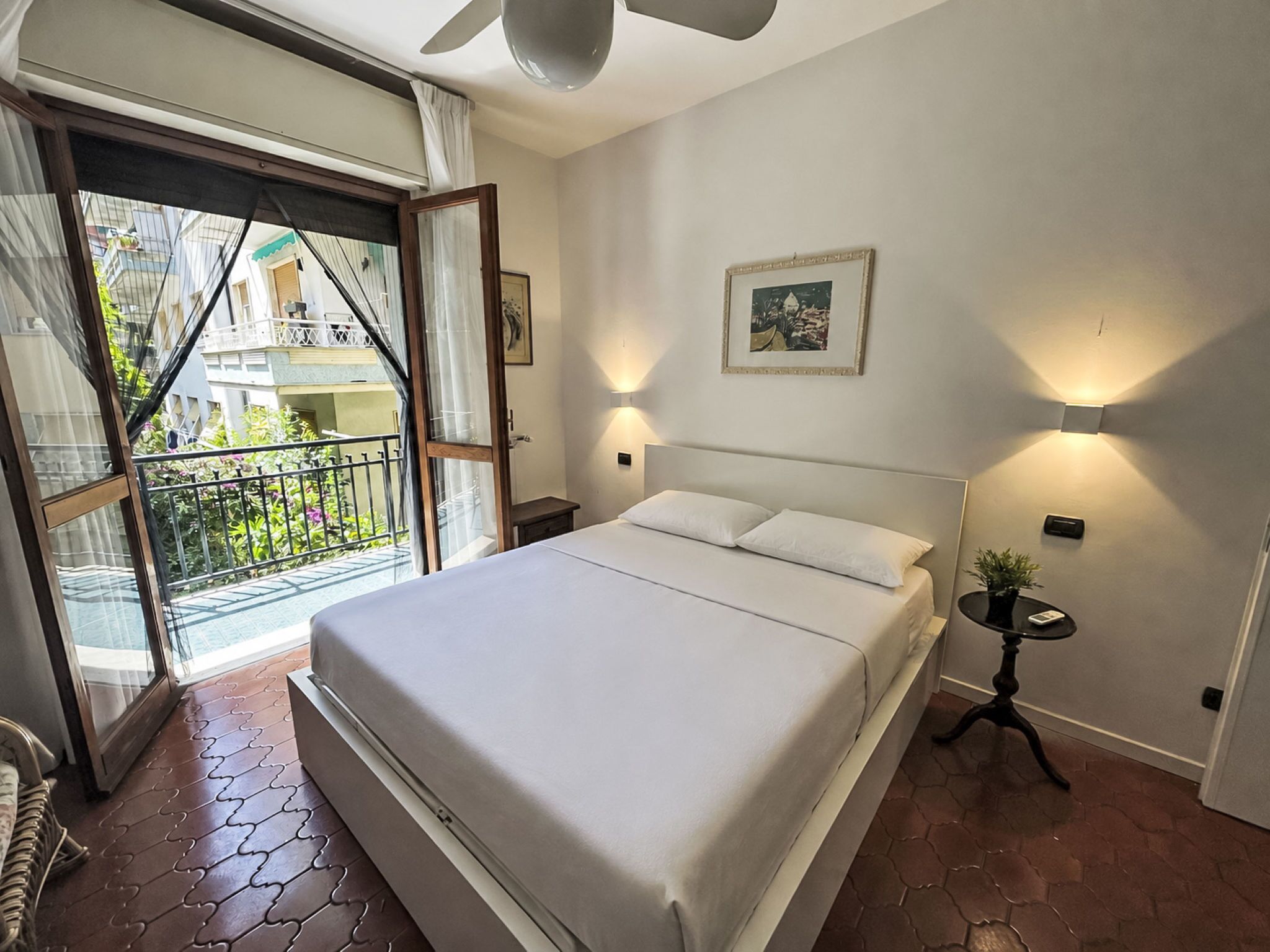 A bedroom with a bed, a nightstand, a wall-mounted lamp, a framed picture, and a balcony with a view of buildings.