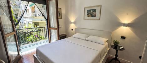 A bedroom with a bed, a nightstand, a wall-mounted lamp, a framed picture, and a balcony with a view of buildings.