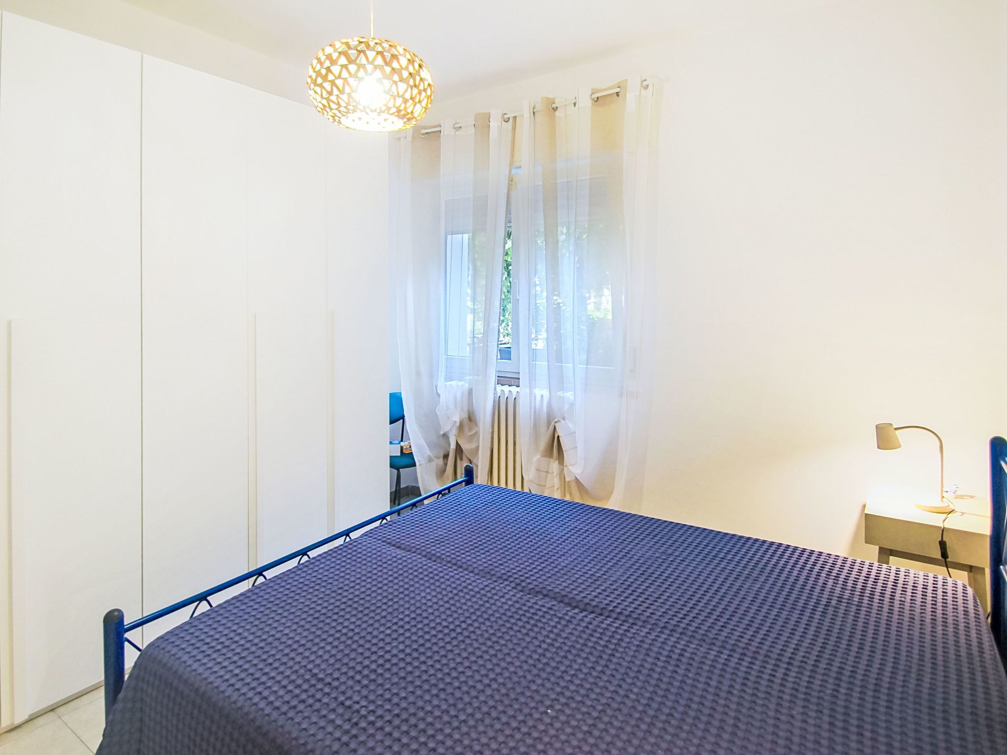 A bedroom with a bed, a desk, a chair, a window with curtains, and a hanging light fixture.