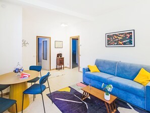 Una sala de estar moderna con un sofá azul, una mesa de comedor con sillas y una mesa de centro. Hay un cuadro en la pared y una planta pequeña sobre la mesa de centro.