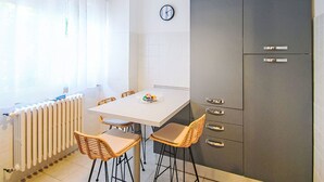 Una cocina compacta con una mesa blanca, sillas de mimbre, un armario gris con cajones, un reloj en la pared y un radiador.