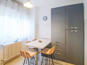 A compact kitchen with a white table, wicker chairs, a grey cabinet with drawers, a clock on the wall, and a radiator. - Boscolago by Interhome (Porto Ceresio)