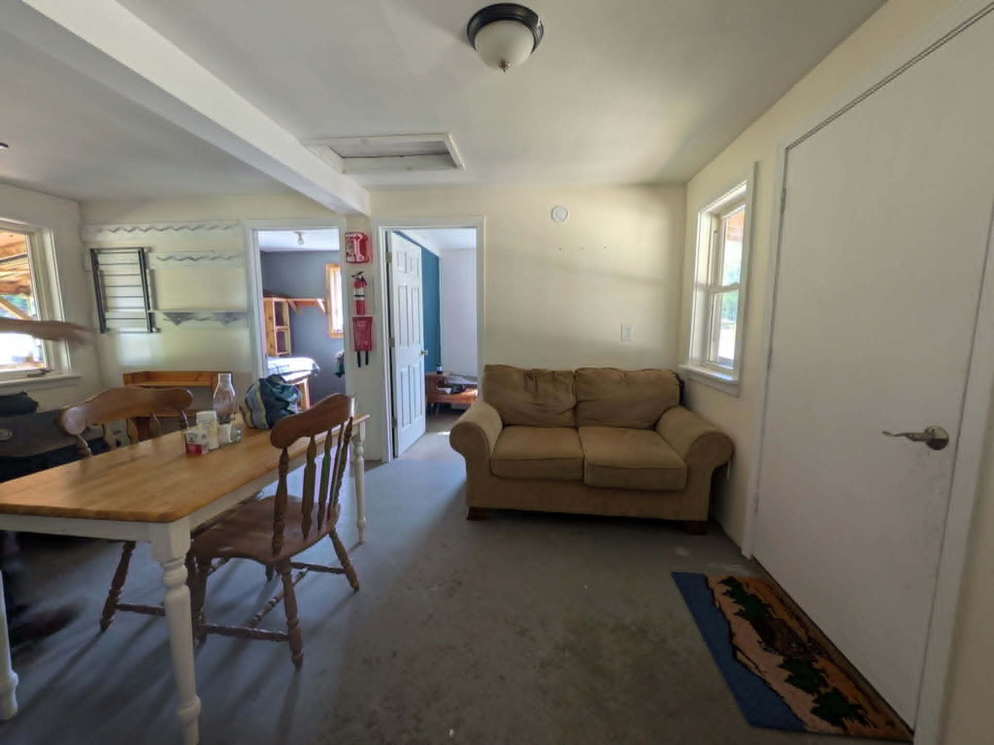A living room with a beige sofa, a wooden dining table with chairs, and a kitchen area in the background.