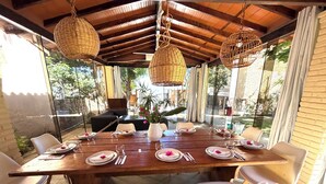 A dining area with a wooden table set for four, surrounded by white chairs, under a wooden ceiling with hanging wicker lamps.