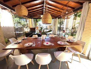 Comedor con una mesa de madera dispuesta para cuatro personas, rodeada de sillas blancas, bajo un techo de madera con lámparas de mimbre colgantes.