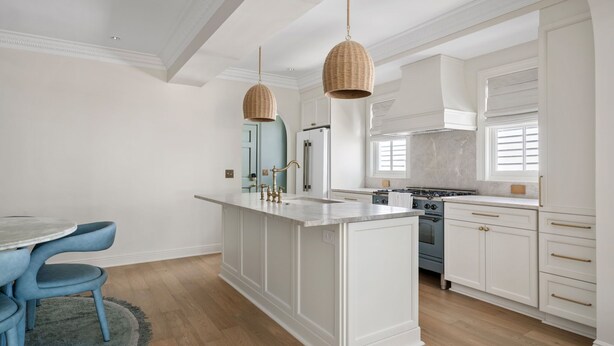 Une cuisine moderne avec des meubles blancs, un îlot central, des luminaires suspendus et un coin repas avec des chaises bleues.