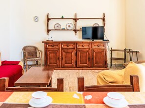 Ein Zimmer mit einem Holzkasten, einem an der Wand befestigten Fernseher, einem Korbstuhl, einem roten Bett und einem Esstisch mit Tellern und Tassen.