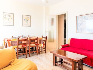 Ein Wohnzimmer mit einem roten Sofa, Holzstühlen, einem Couchtisch aus Holz und einem Esstisch mit Stühlen. An der Wand hängen zwei gerahmte Kunstwerke.