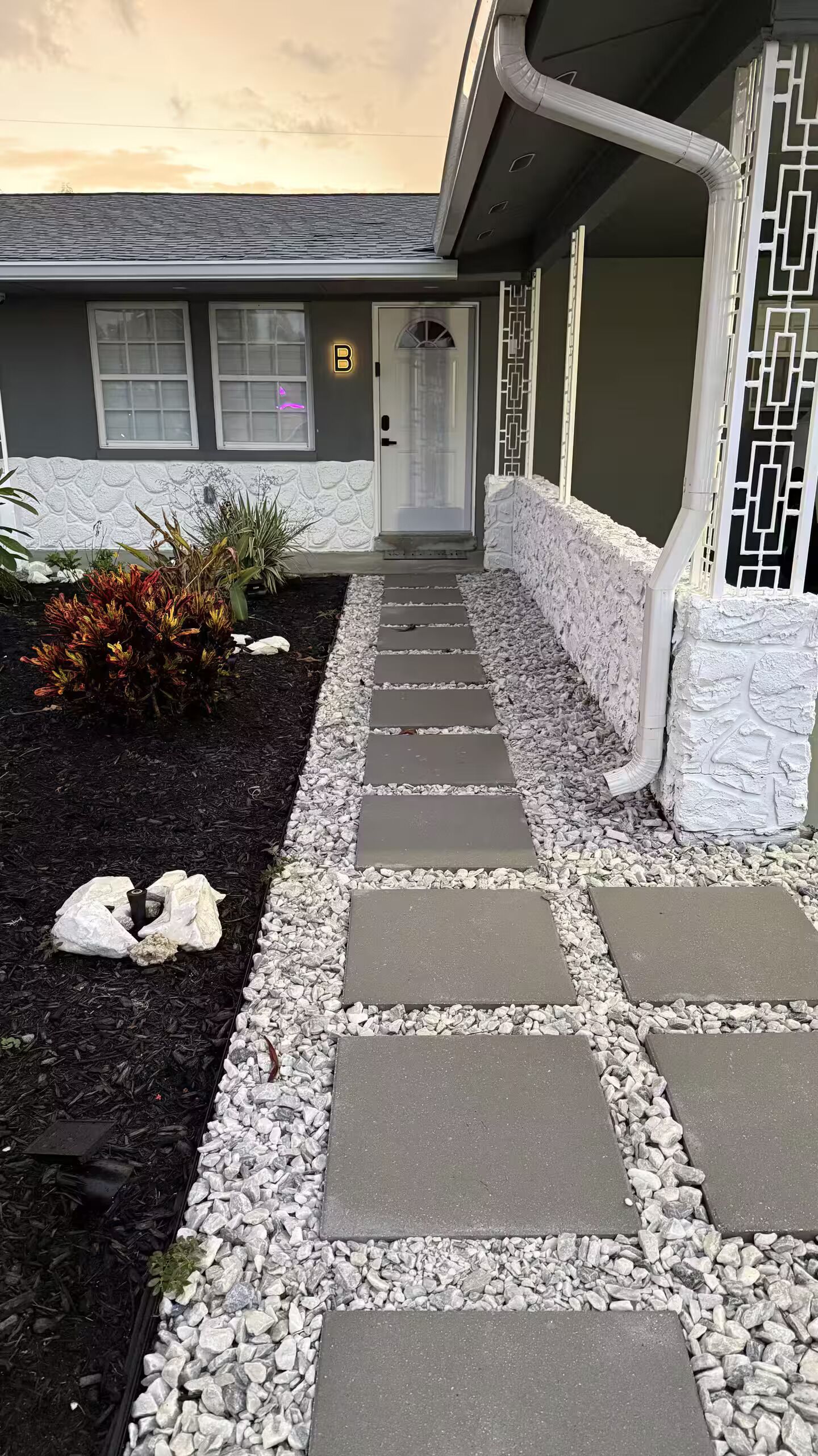 A pathway leading to a house with a grey facade, a white door, and a small garden with plants and stepping stones.