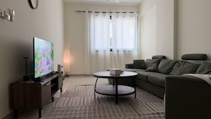 A living room with a flat-screen TV, a sofa, a coffee table, and a lamp. - The Hive - Cozy 2BR Apartment with Rooftop Pool, Al Khuwair Muscat (Muscat)