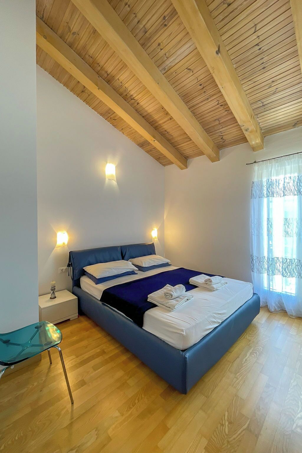 A bedroom with a wooden ceiling, a bed with blue bedding, bedside lamps, and a window with curtains.