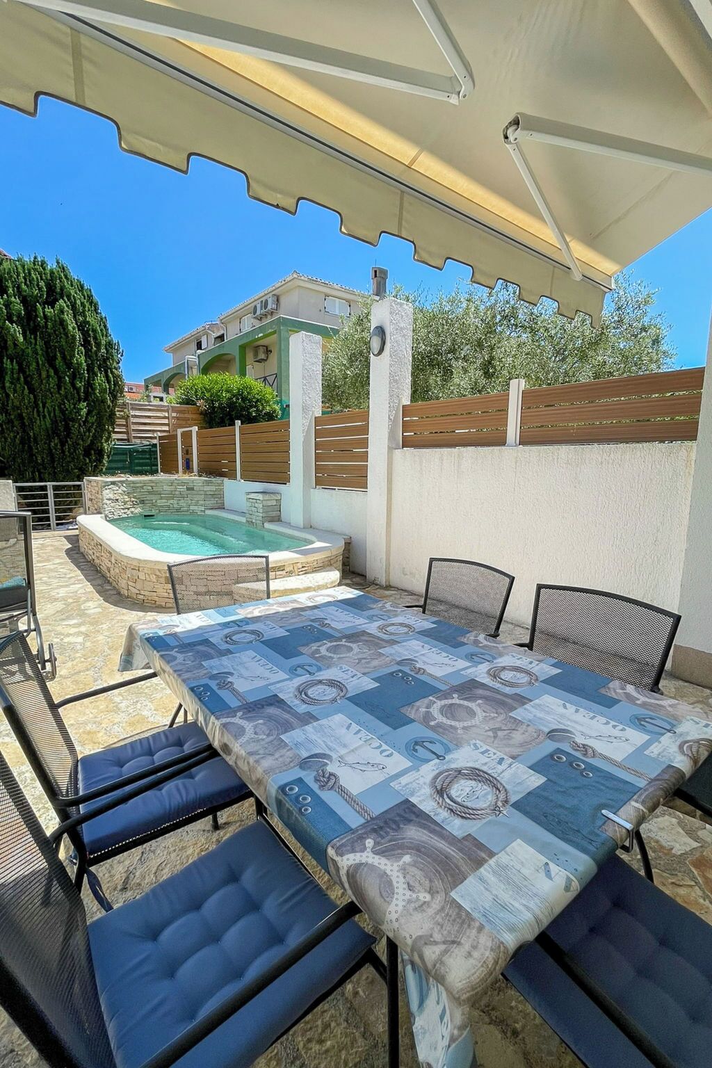 En udendørs patio med pool, et overdækket siddeområde og udsigt til omkringliggende bygninger.