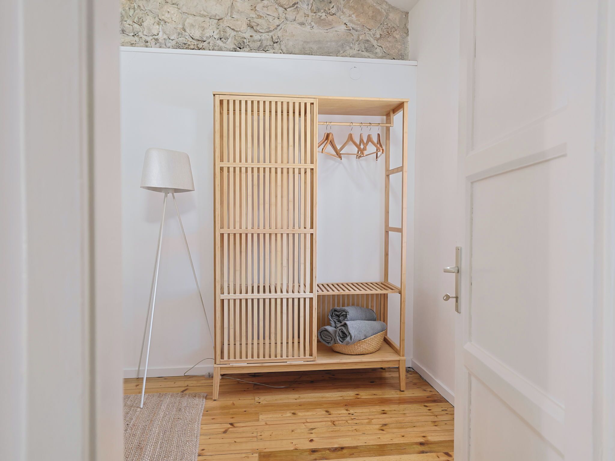 A wooden wardrobe with a hanging rack, a basket with folded clothes, and a floor lamp.
