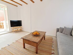 Un salon avec une table basse en bois, un canapé gris, un téléviseur à écran plat posé sur un meuble en bois et un panier d’oranges sur la table.