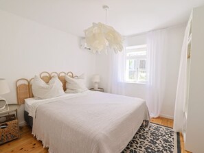 Ein Schlafzimmer mit einem großen Bett, einem Nachttisch, einer Lampe, einem Korbwannenstuhl und einem Fenster mit Vorhängen.
