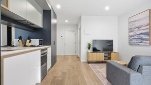 A modern kitchen with white cabinetry, a microwave, and a sink. There is a dining area with a grey sofa and a flat-screen TV on a wooden stand. - Your Melbourne Getaway Base in Carlton (Carlton)