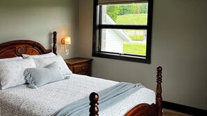 A bedroom with a wooden bed, a nightstand, a window with a view of a green landscape, and an air conditioning unit. - Charming Farmhouse on a Cherry Orchard just a few minutes from the beach. (Suttons Bay)