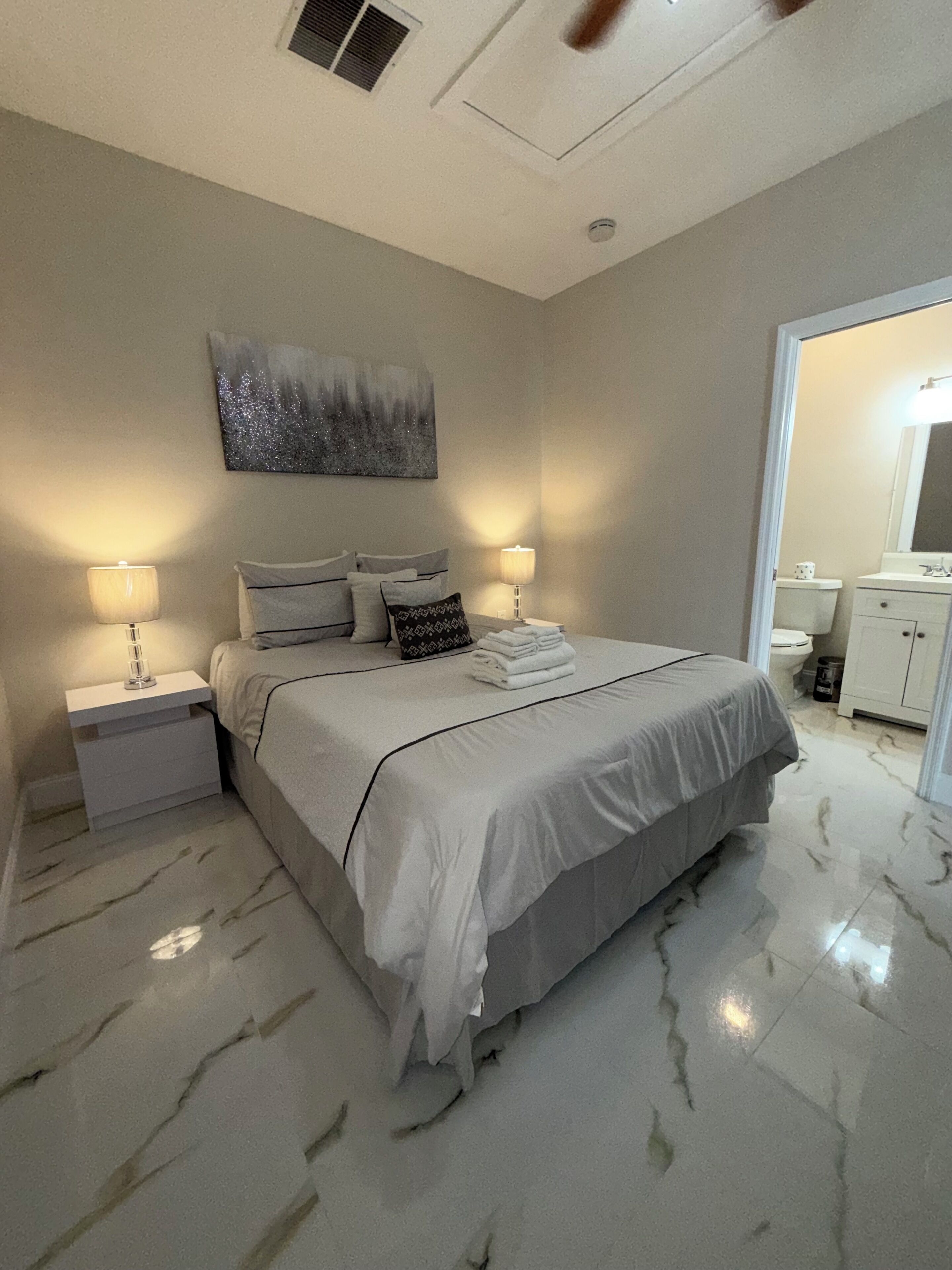 A bedroom with a bed, nightstands, a painting, and a bathroom visible through an open door.