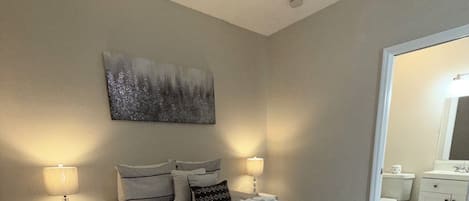 A bedroom with a bed, nightstands, a painting, and a bathroom visible through an open door.