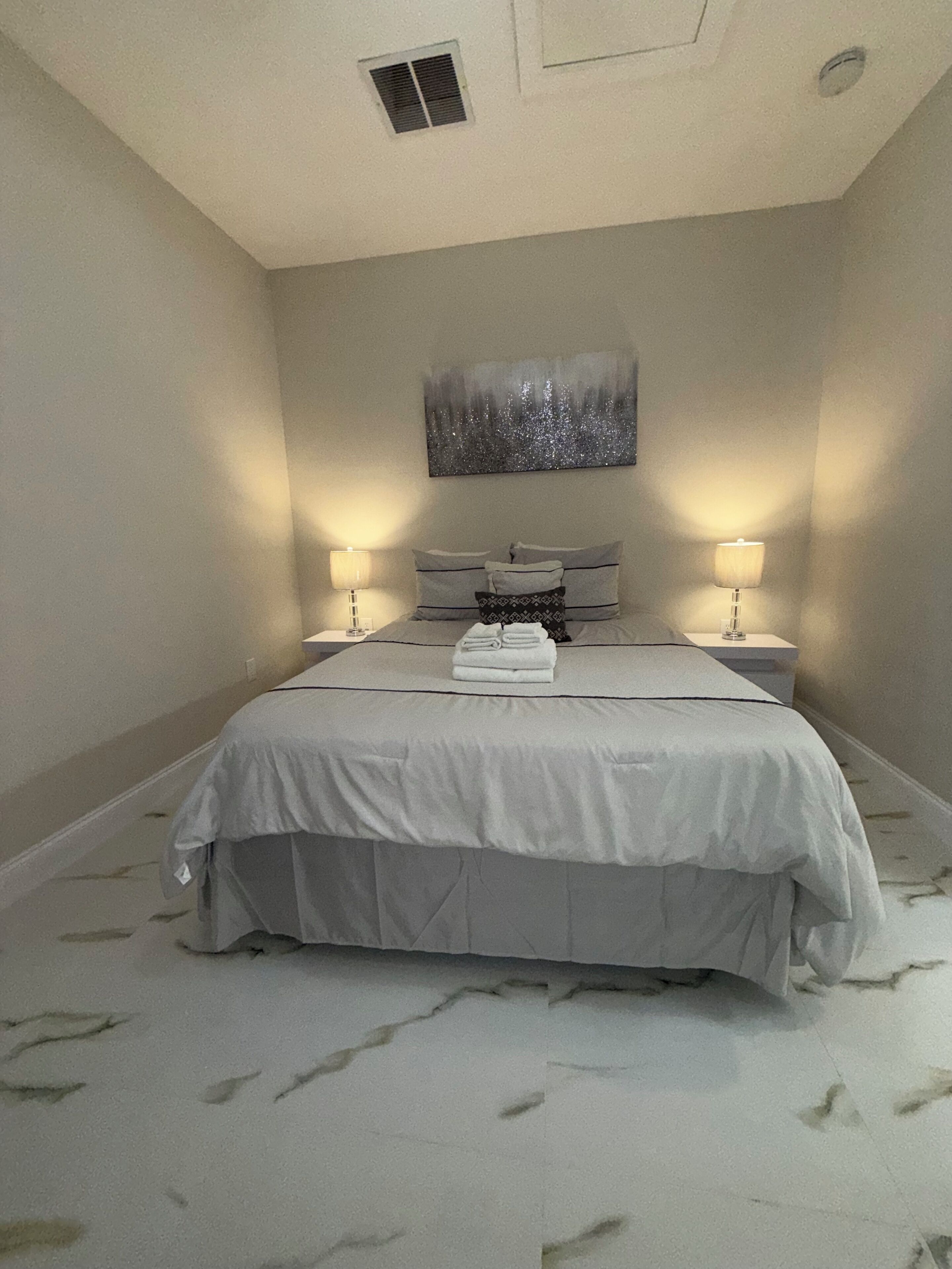 A bedroom with a bed, two bedside lamps, a painting on the wall, and a ceiling vent.
