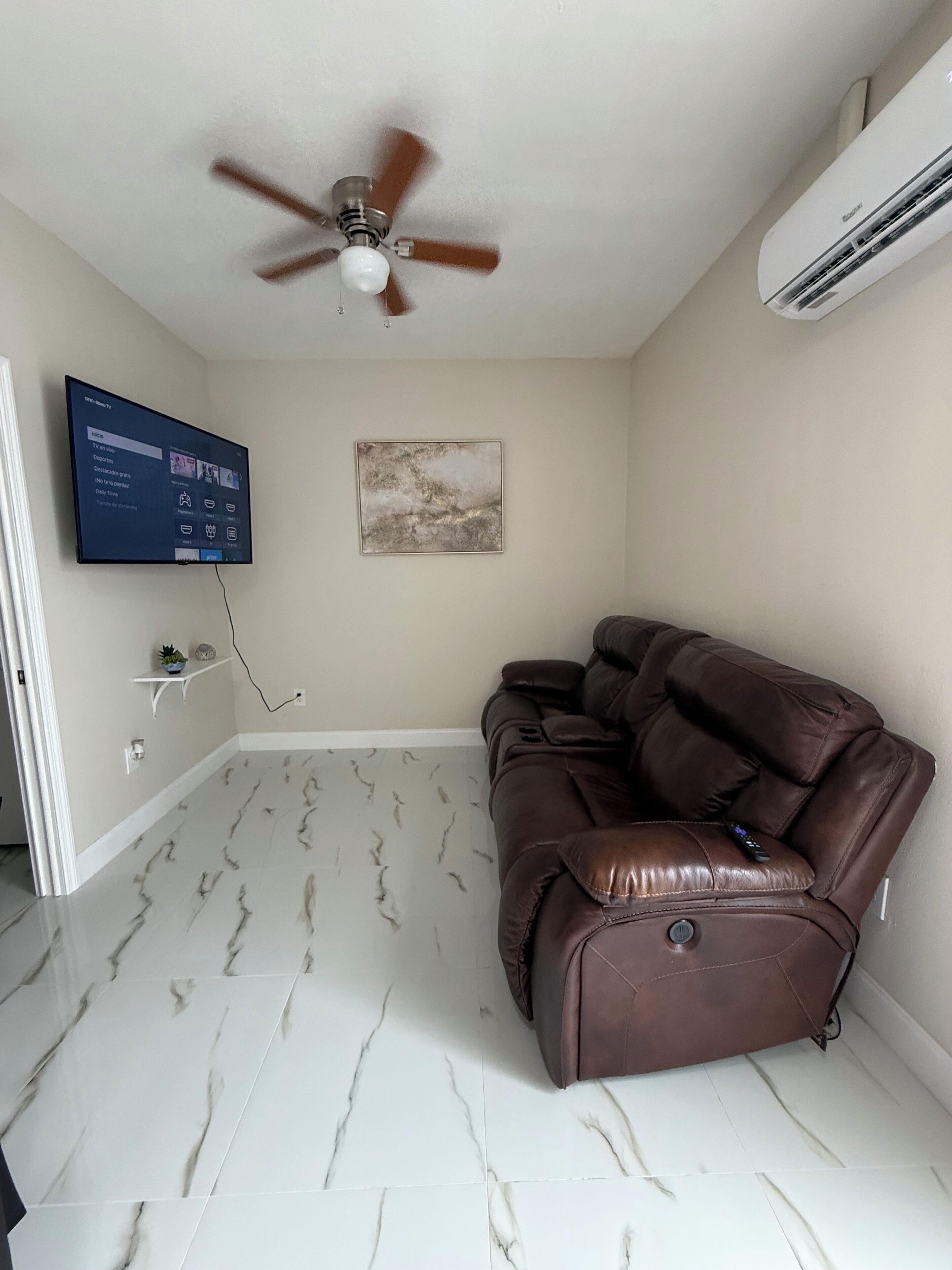 A room with a ceiling fan, a mounted flat-screen TV, a brown leather recliner, and a marble-patterned floor.