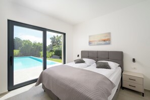 Ein Schlafzimmer mit einem Bett, einem Nachttisch, einem Gemälde und einer Glasschiebetür, die zu einem Pool führt.