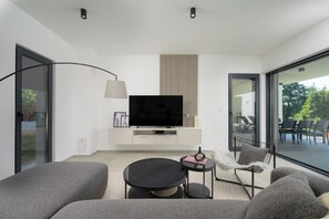 Ein modernes Wohnzimmer mit einem Flachbildfernseher, grauen Sofas, einem kleinen runden Tisch und einem Beistelltisch mit einer Lampe.