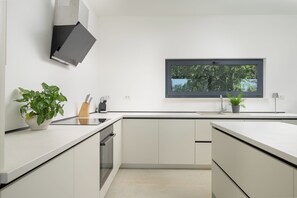 Eine moderne Küche mit weißen Schränken, einem eingebauten Ofen, einem Fenster mit Blick auf Bäume und einem Topf mit einer Pflanze auf der Arbeitsfläche.