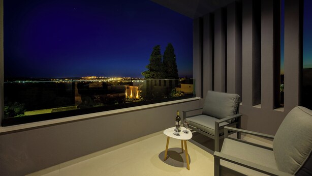 Uma varanda com vista para a paisagem urbana noturna, uma mesa com garrafa e copo, e uma cadeira.