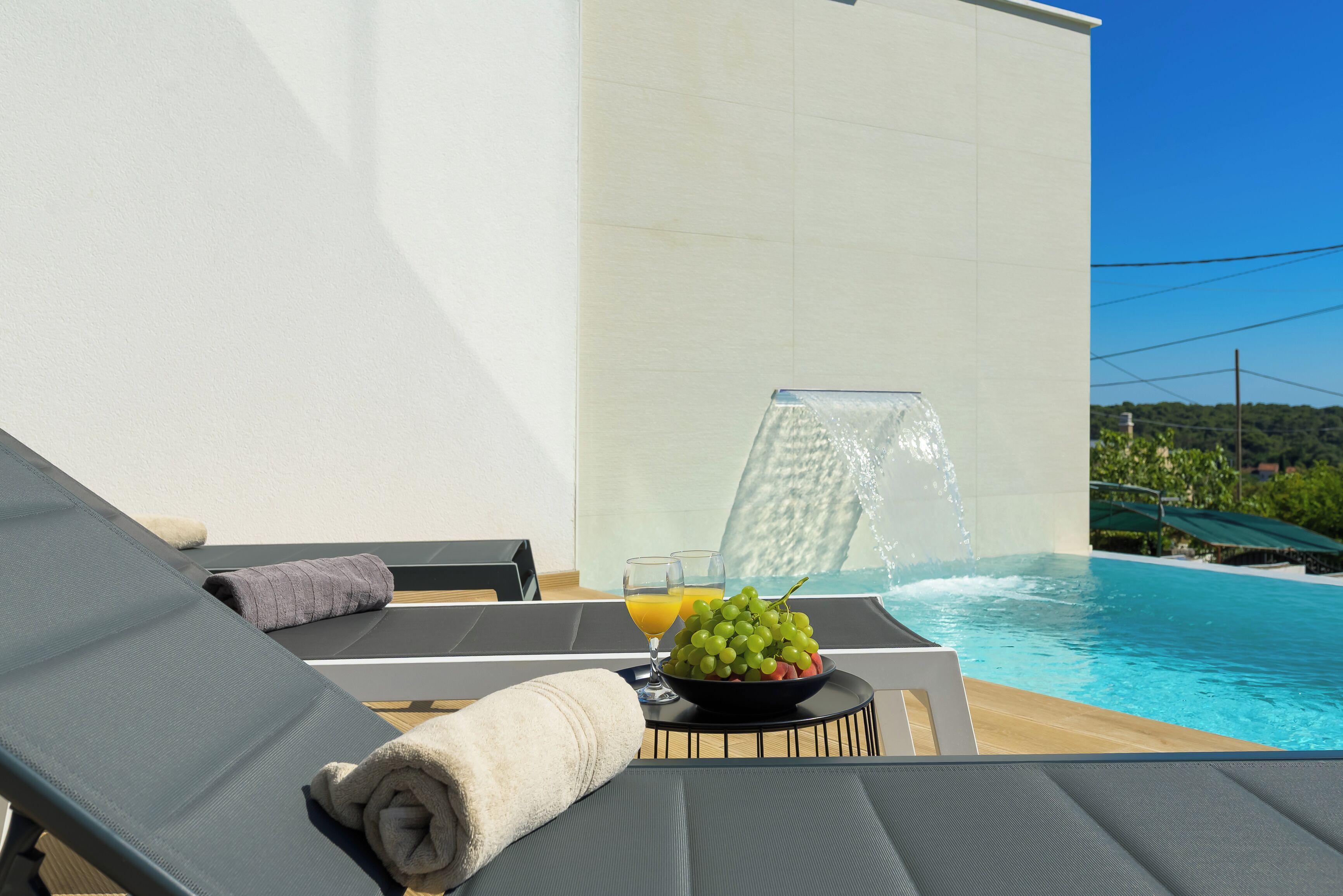 A poolside lounge with a water feature, a bowl of fruit, and a glass of orange juice.