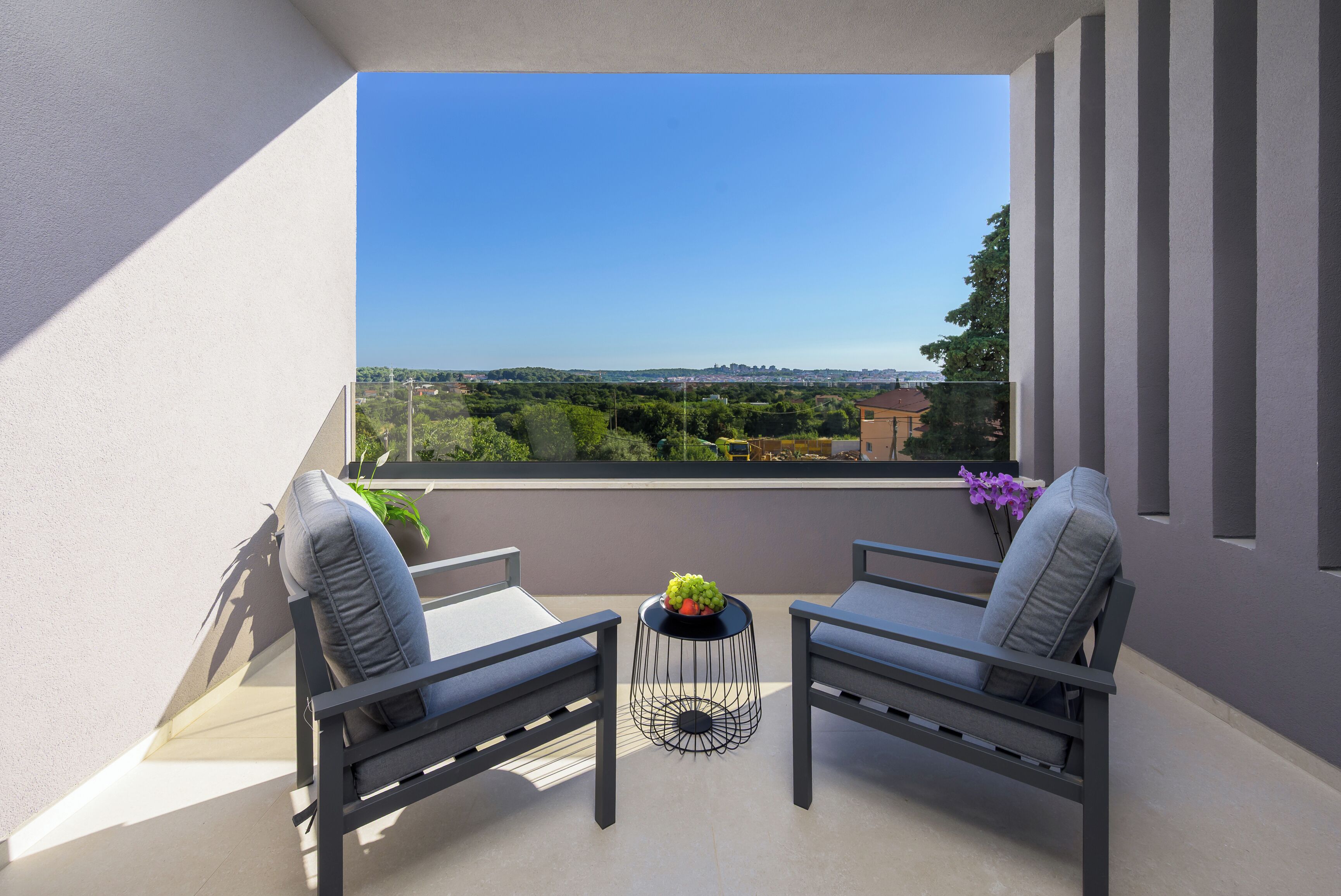 A balcony with two chairs, a small table with a fruit bowl, and a view of a cityscape.