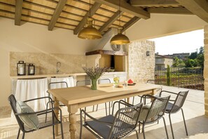 An outdoor dining area with a stone wall, a wooden table, metal chairs, and hanging pendant lights.