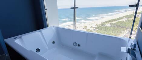 A modern bathroom with a large bathtub, a window with a view of the beach, and a glass partition.