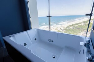 A modern bathroom with a large bathtub, a window with a view of the beach, and a glass partition.