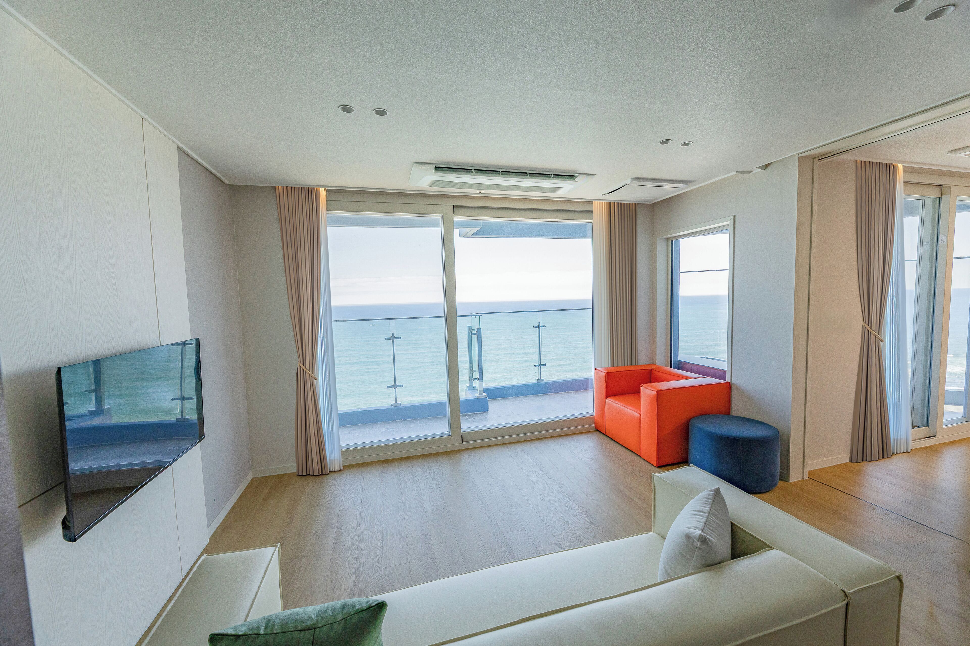 Ein modernes Wohnzimmer mit Flachbildfernseher, einer Couch, einem kleinen orangefarbenen Sessel, einem blauen Hocker und großen Schiebeglasüren zum Balkon mit Meerblick.