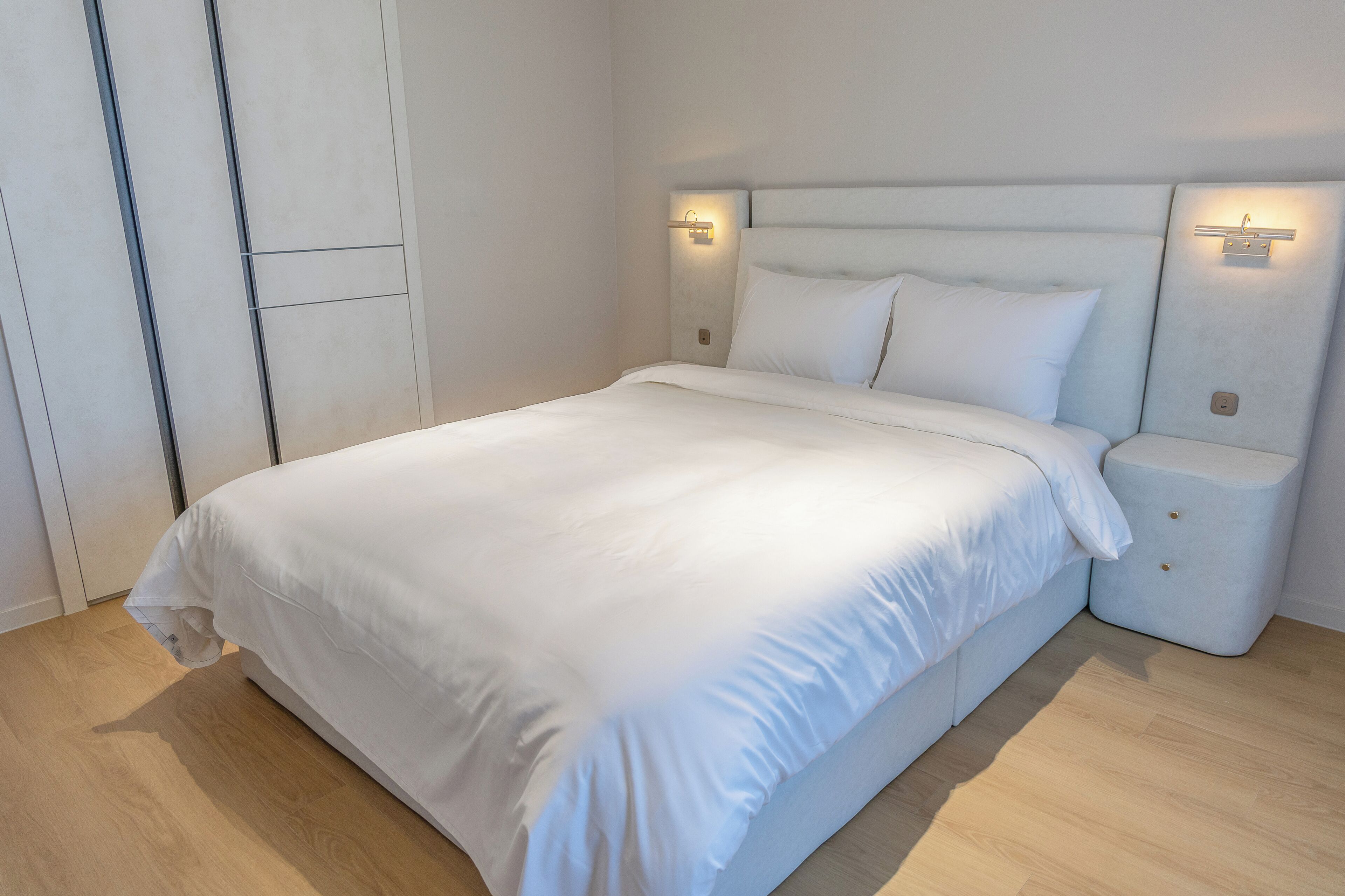 A neatly made bed with white linens, a light-colored headboard, and bedside lamps.