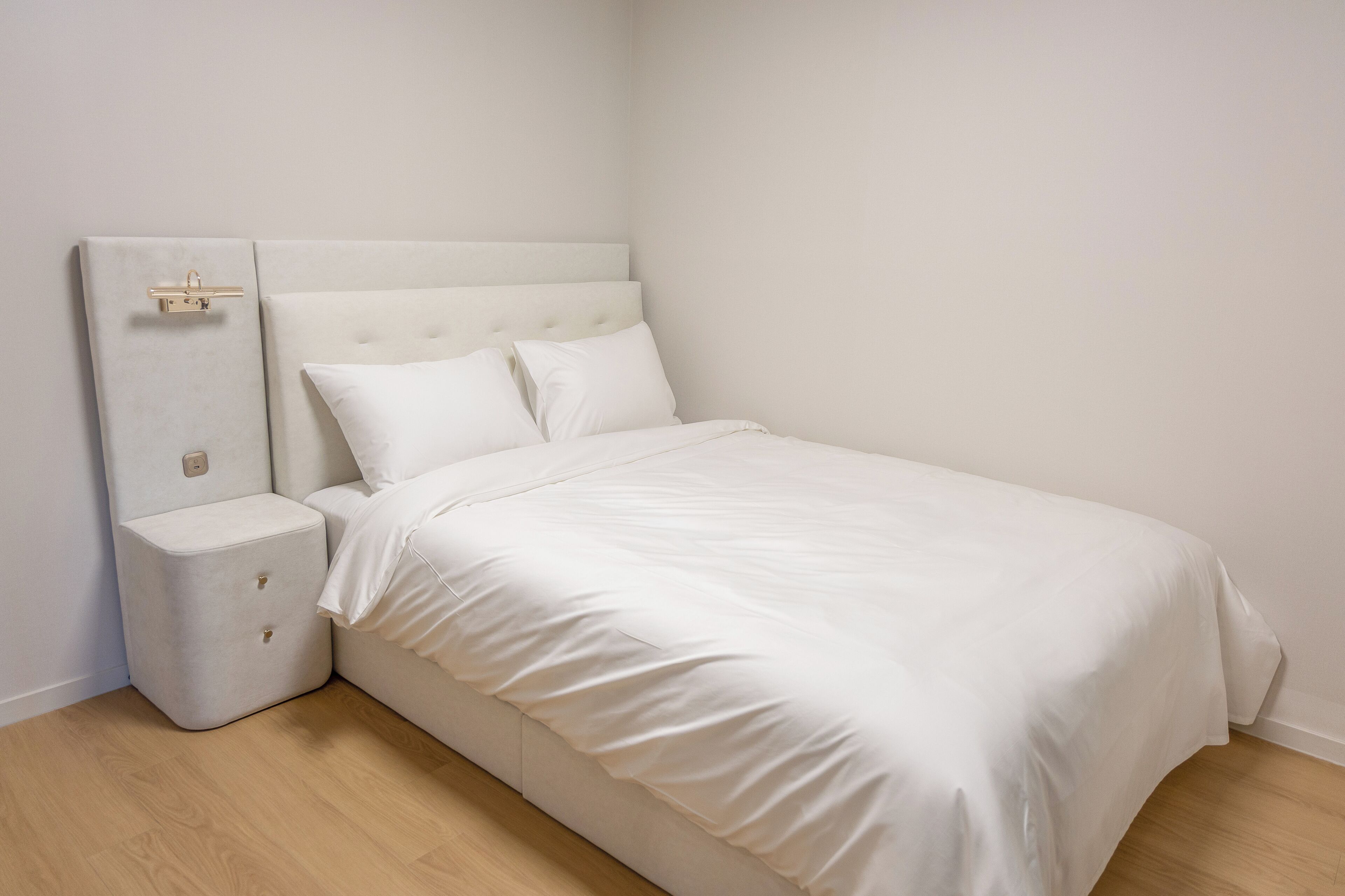 A neatly made bed with a headboard, a bedside table with drawers, and a wall-mounted light fixture.