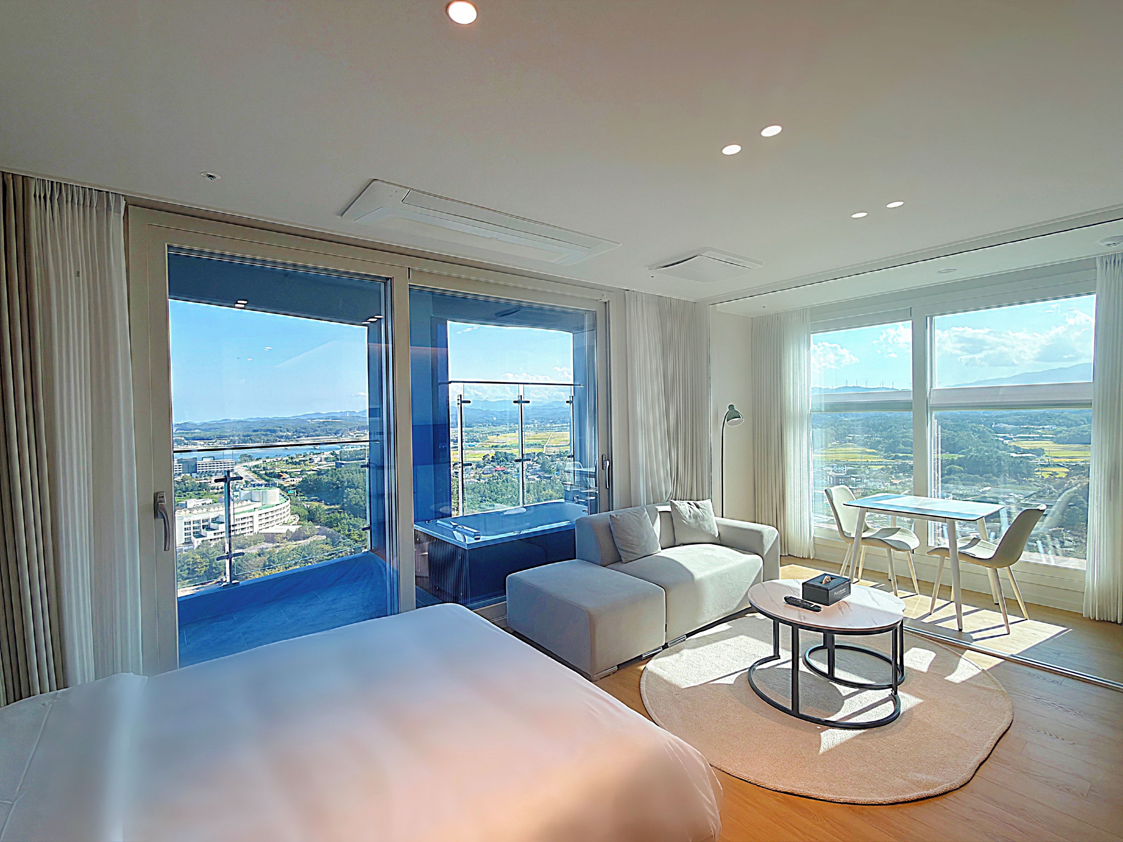 Ein modernes Hotelzimmer mit einem großen Bett, einer Sitzecke, einem kleinen Tisch und Blick auf eine Stadtlandschaft durch große Fenster.