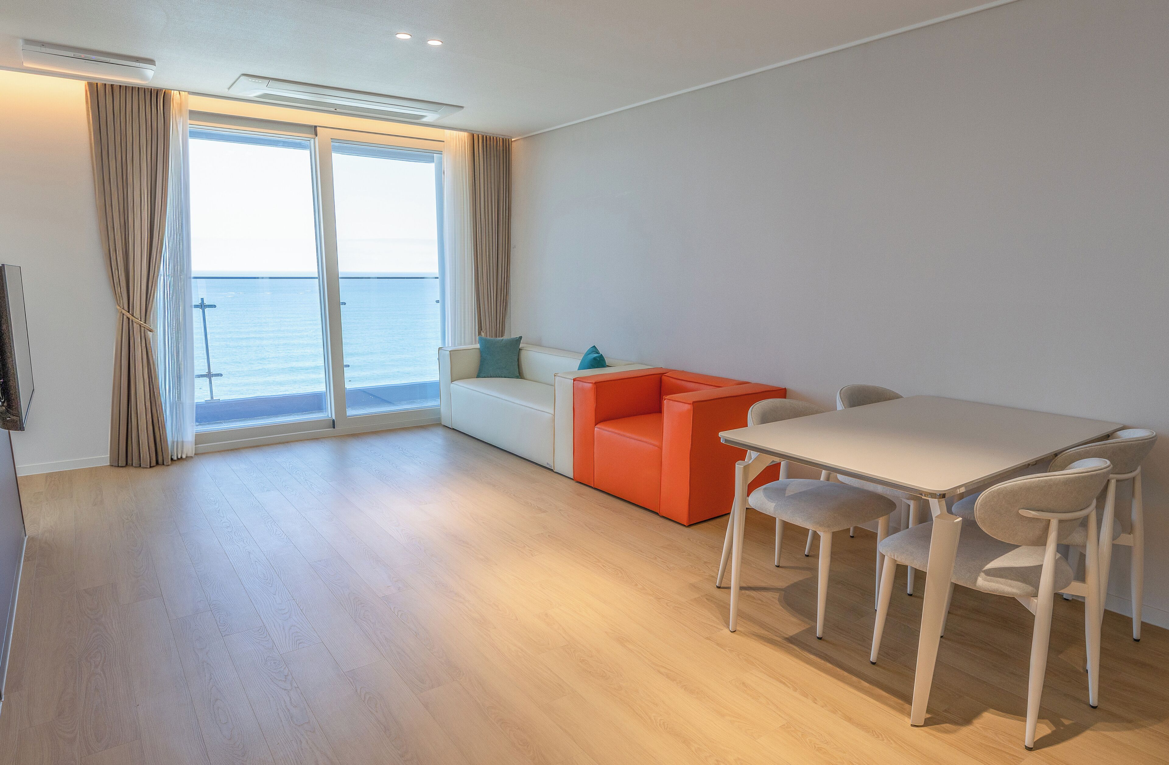 A modern room with a wooden floor, a white table with four chairs, a red sofa, and a large window with curtains.