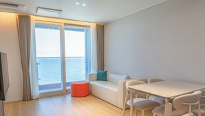 Una habitación moderna con un sofá blanco, una mesita pequeña y sillas, un reposapiés rojo y vistas al mar a través de amplios ventanales.