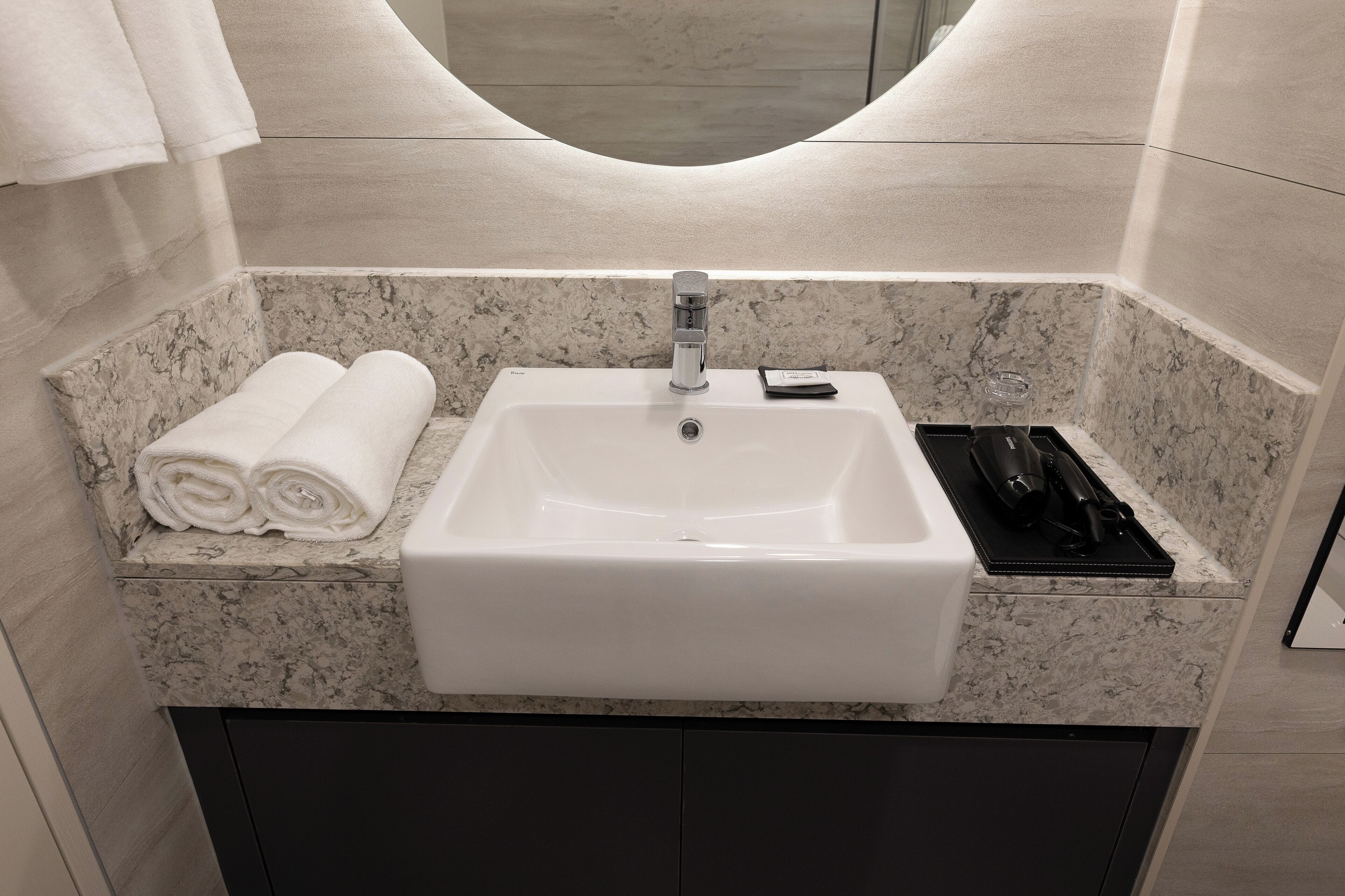 A bathroom with a white sink, a towel rack with folded towels, and a black tray with toiletries.