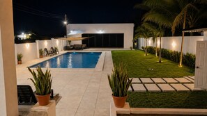 Una piscina con zona de terraza, césped y un edificio blanco con porche techado.